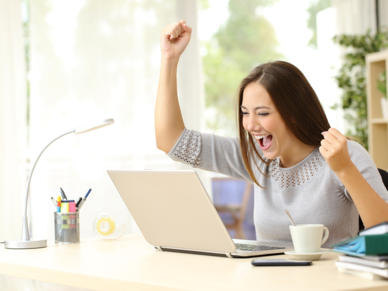 A woman smiles and raises her arms while looking at a computer