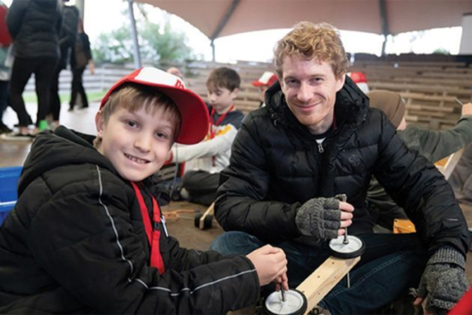 A young student poses with a teacher while they fix a go-kart